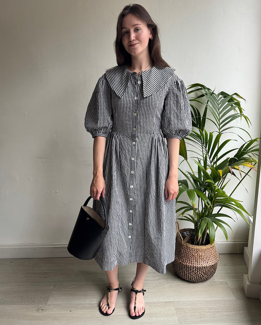 A woman standing in a room with a plant backdrop, wearing a black and white gingham seersucker shirt dress with a statement collar, button fastenings, elbow length sleeves, and pockets. She is also carrying a Longchamp leather bucket bag and wearing black leather sandals with gold beads.