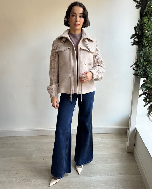 A woman wearing a cream-colored jacket with a knitted sleeve and a drawstring waist, paired with blue jeans. She is also wearing a beige knit sweater and white platform shoes.